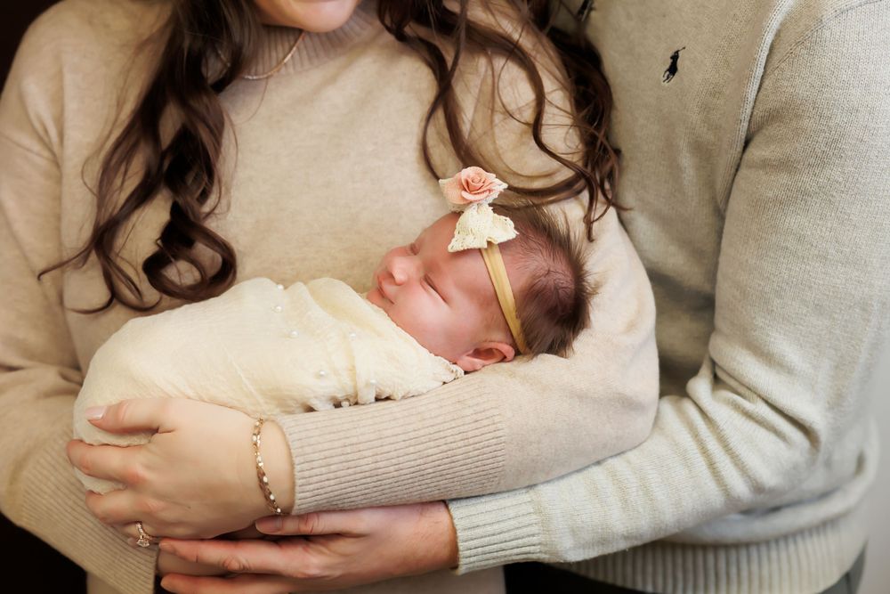 Tender family moment with newborn baby wearing bow headband cradled in warm sweater-clad arms.