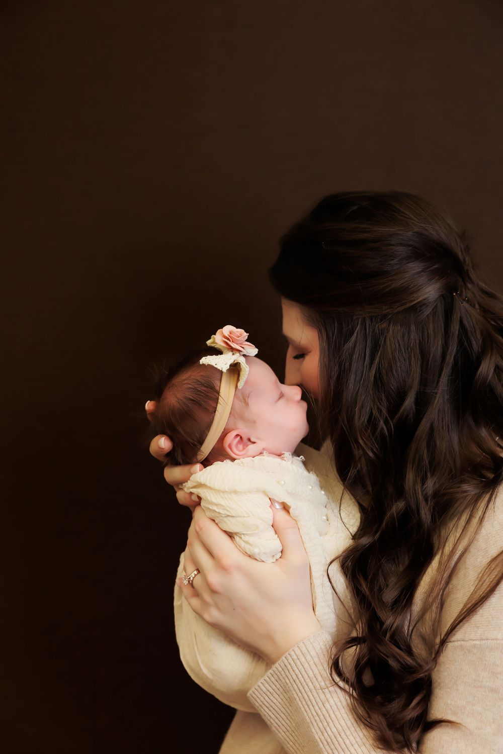 Mother in beige sweater lovingly kisses baby against dark brown backdrop.