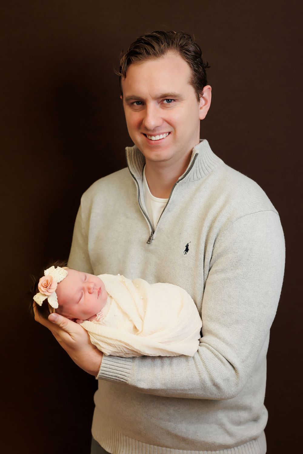 Person in gray sweater holding sleeping newborn baby in cream-colored wrap against brown background.