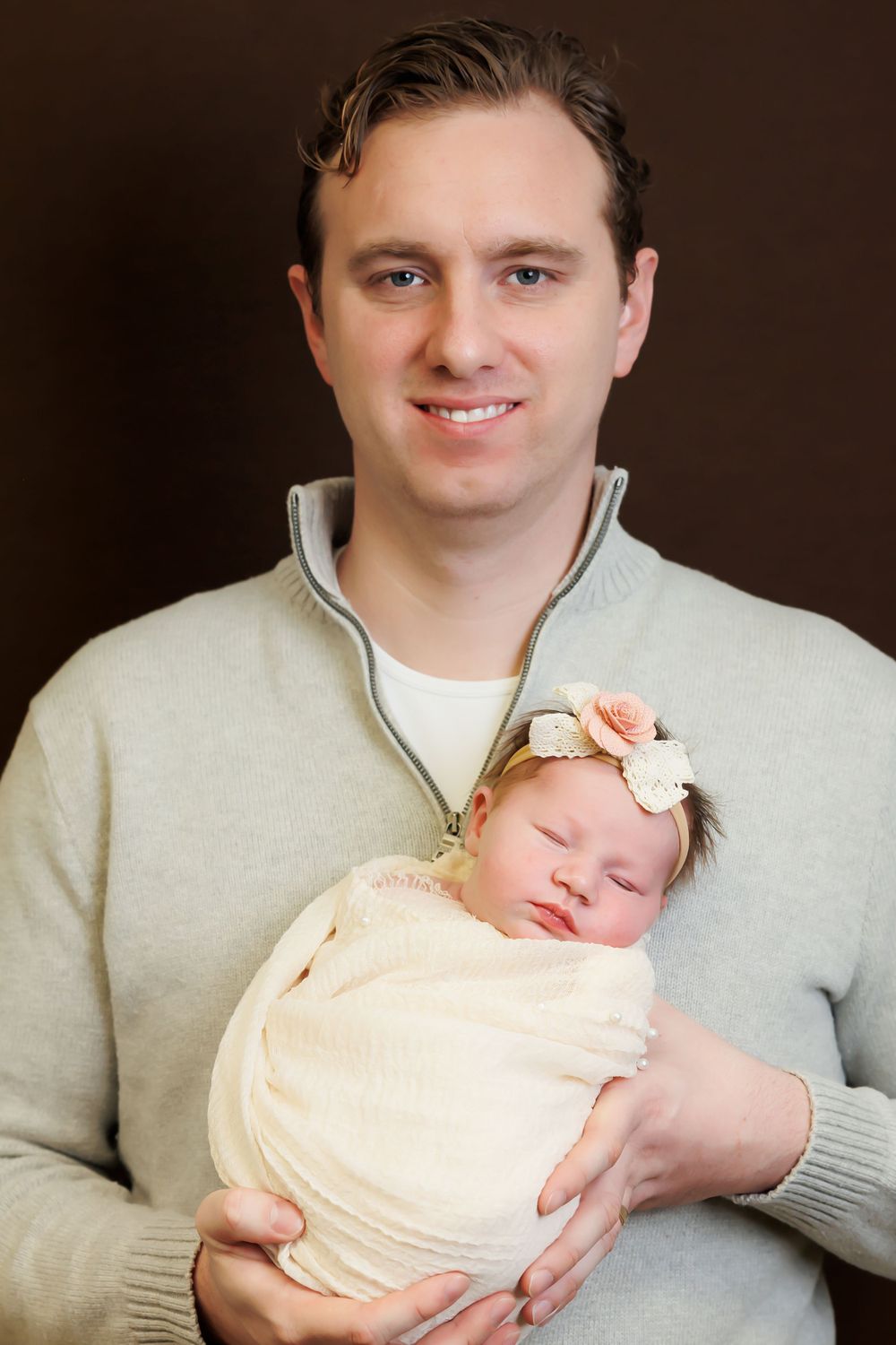 Portrait of parent in grey sweater holding sleeping newborn wrapped in cream fabric.
