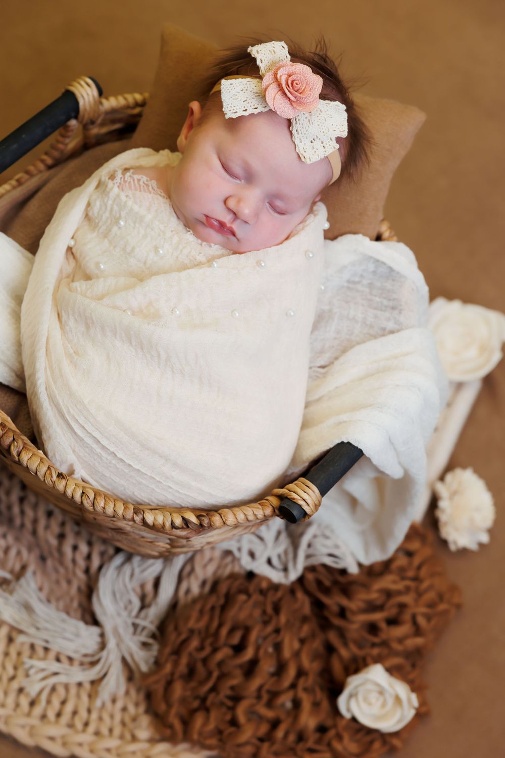 Sleeping newborn wrapped in cream fabric with pink flower headband rests in rustic woven basket with knit textiles.