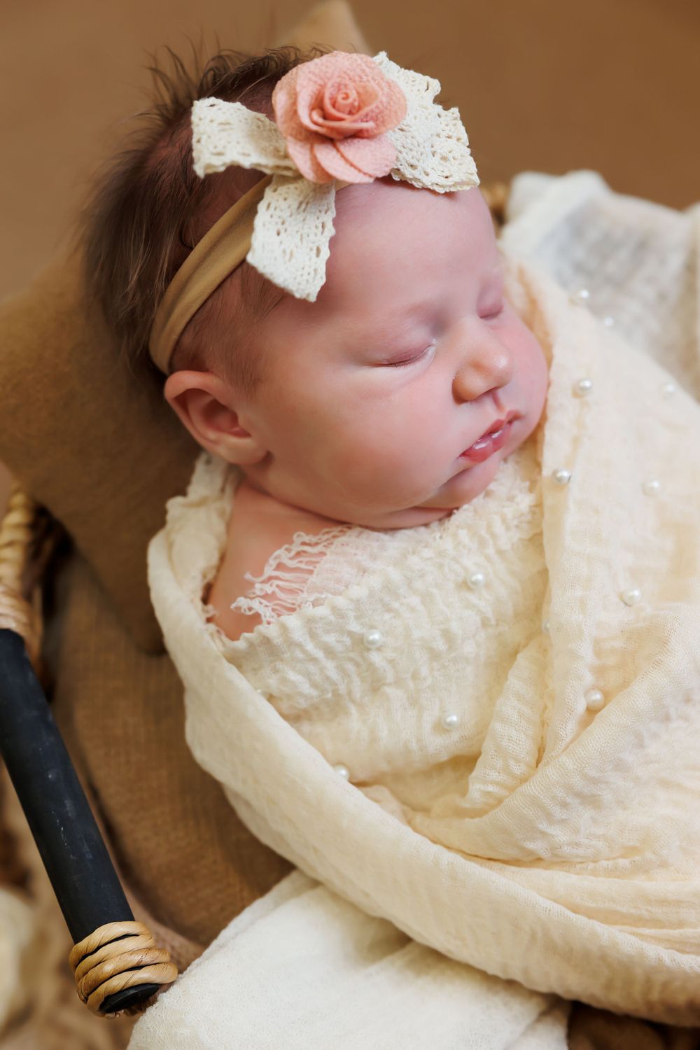Sleeping baby wearing cream lace dress and pink flower headband with white lace bow rests peacefully in soft lighting.