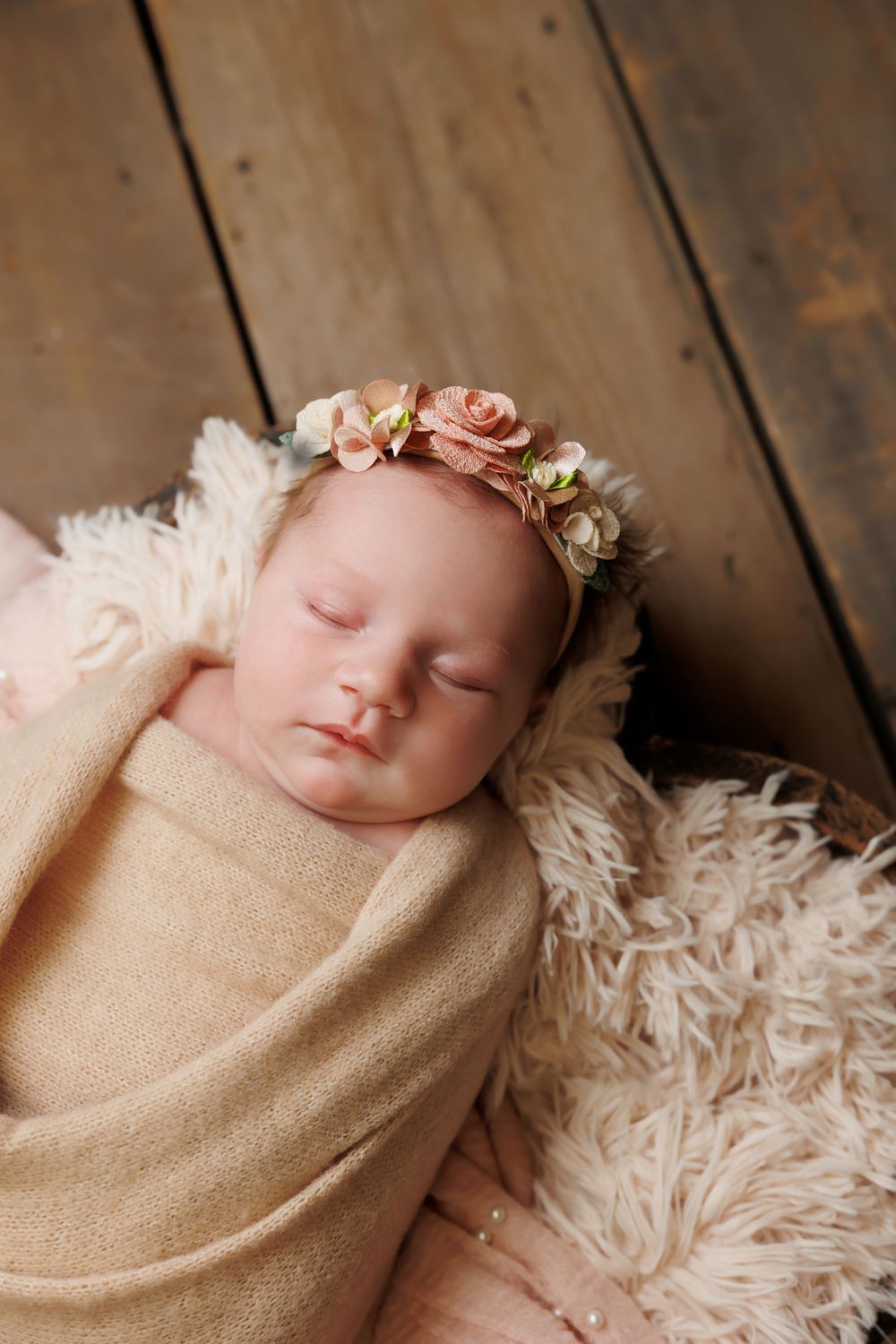 Sleeping newborn wrapped in tan swaddle with floral headband rests on fluffy white fur backdrop.