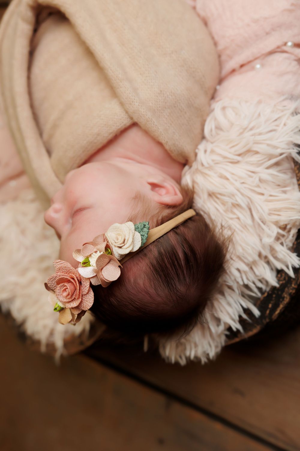 Sleeping newborn swaddled in neutral tones with delicate floral headband on white furry backdrop.