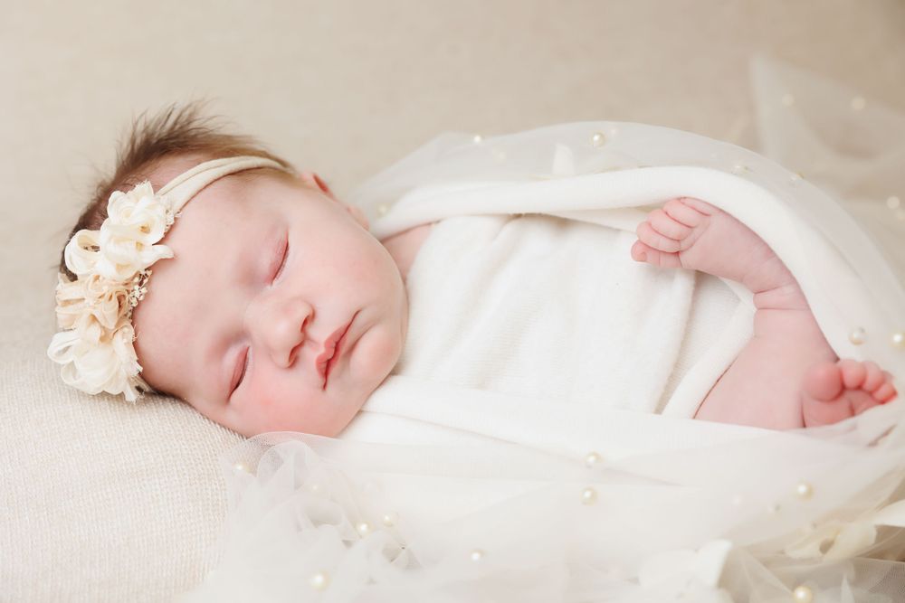 Peaceful sleeping infant wrapped in soft white fabric and wearing a delicate floral headband.