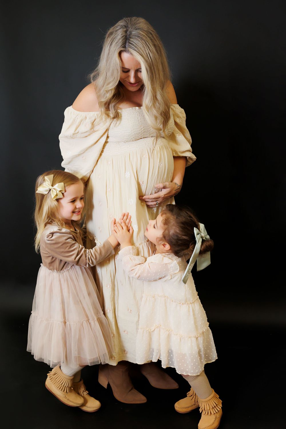 A pregnant woman in cream dress stands with two young children touching her belly in a dark studio setting.