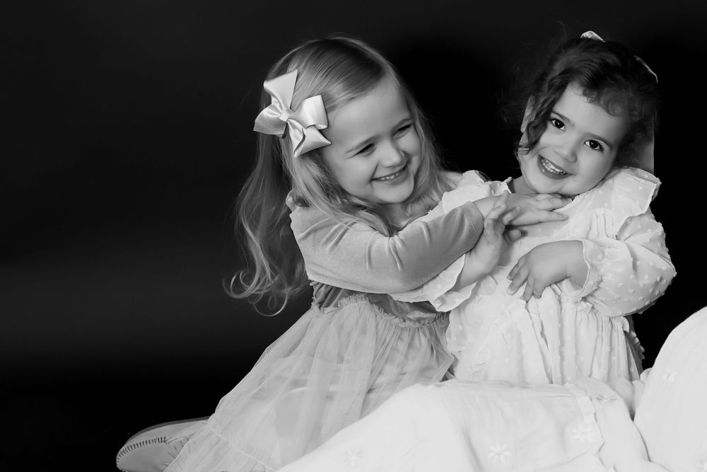 Black and white portrait of two children in matching dresses sharing playful moment.