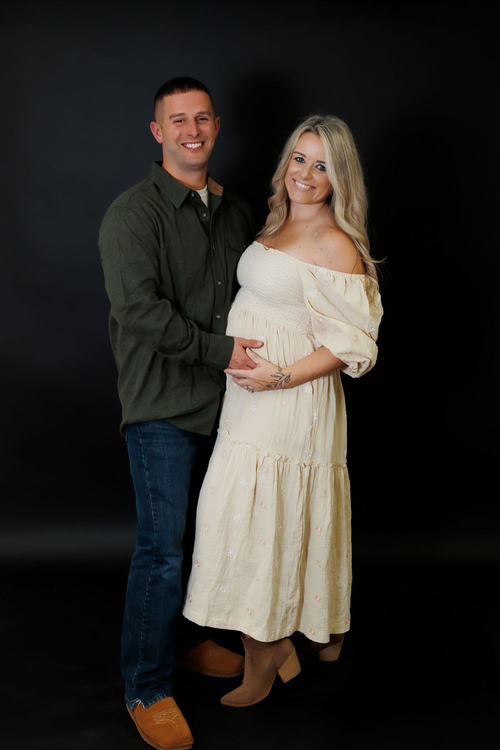 A couple poses together for a maternity photo against a dark background, wearing casual and white dress attire.