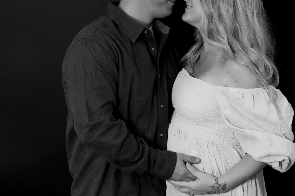 Black and white maternity photo showing an intimate couple's moment with woman in white dress and partner in dark shirt.