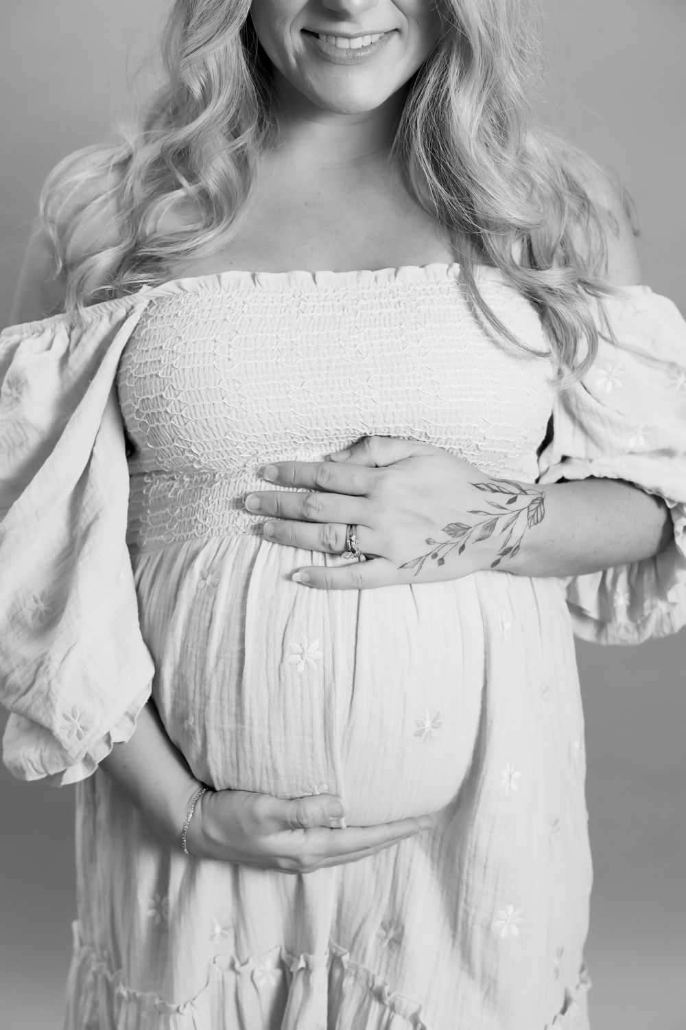 A black and white photo of a pregnant woman in a white off-shoulder dress holding her baby bump.