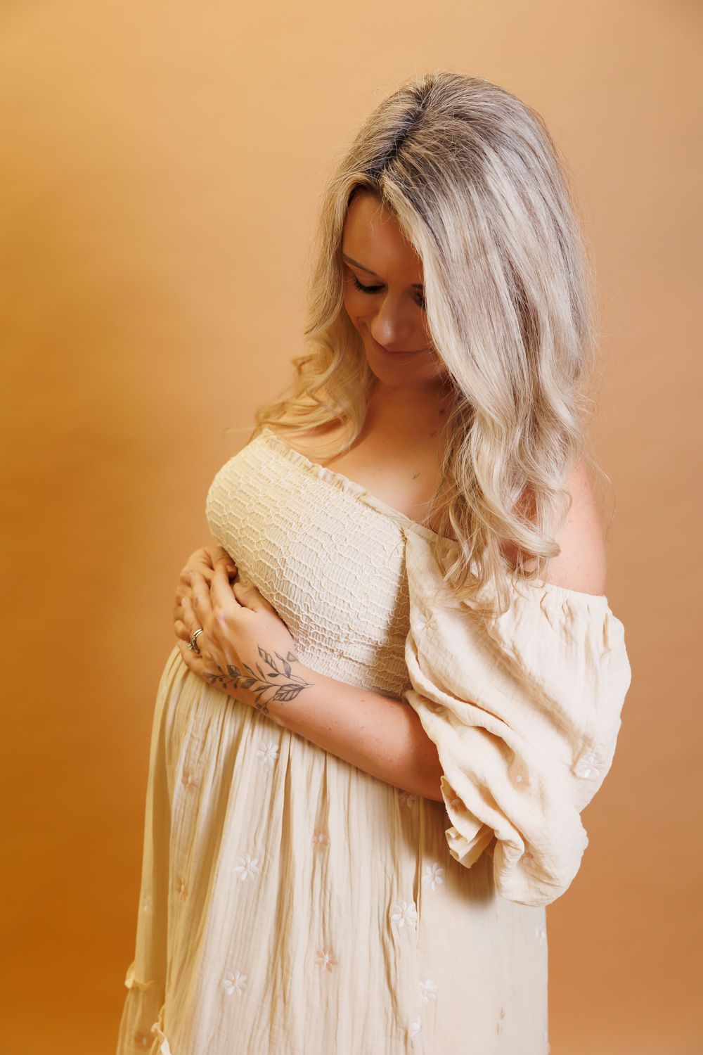 Woman in flowing cream off-shoulder dress poses against warm background during maternity photoshoot.
