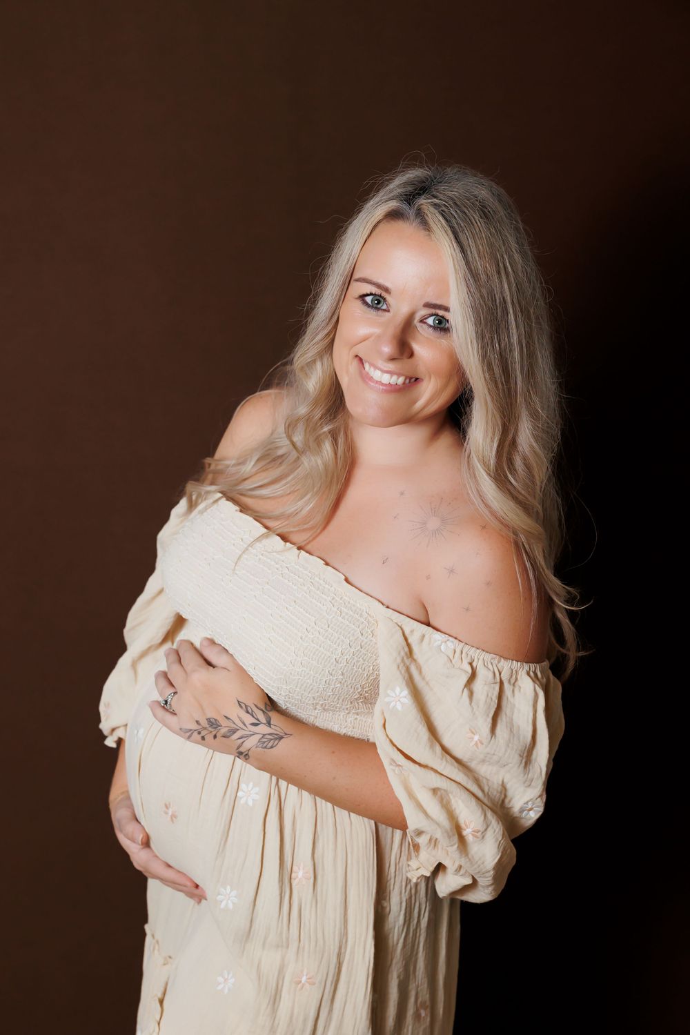 Expectant mother in cream off-shoulder dress smiles during studio maternity portrait session.