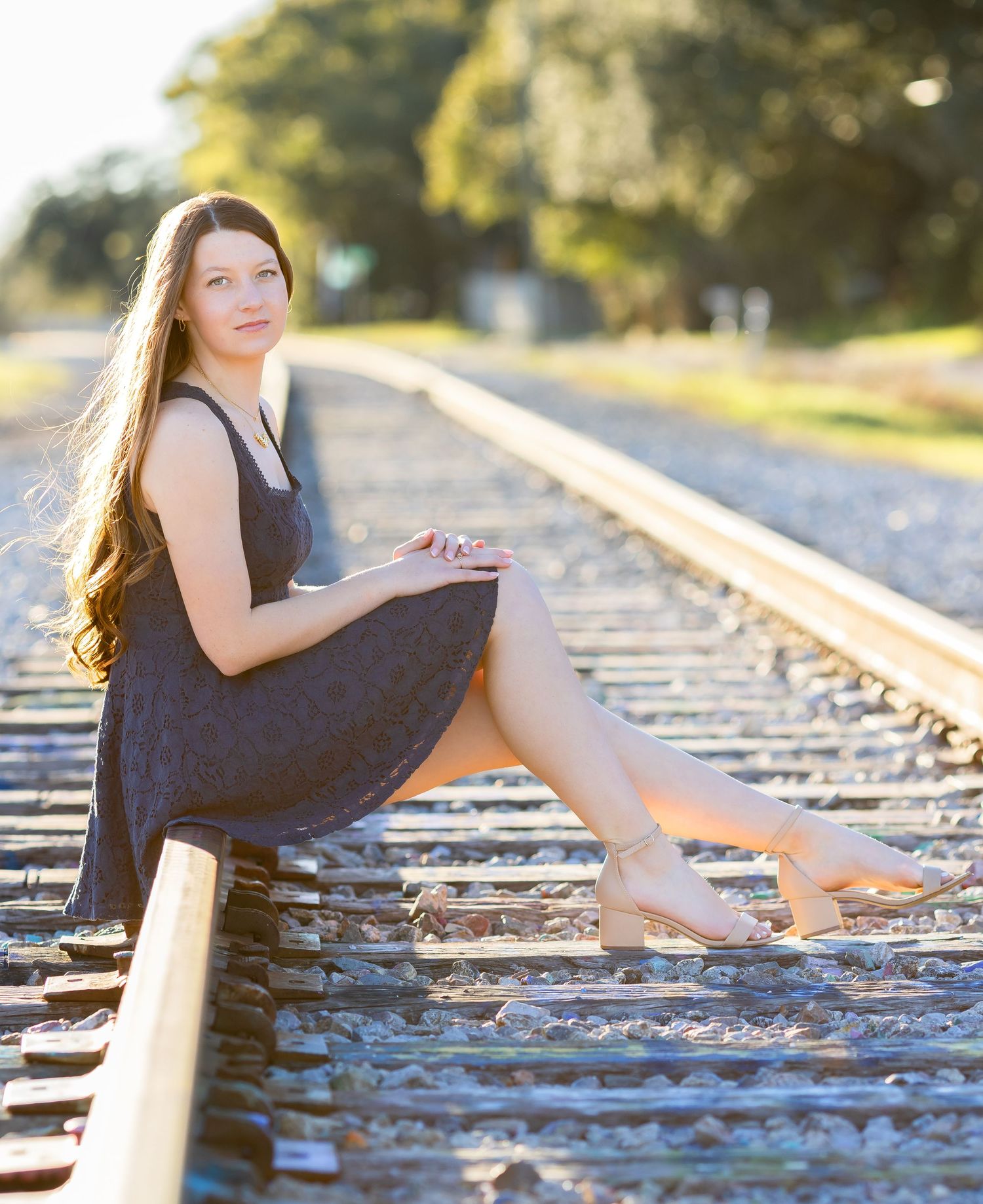 Person in a black dress sitting on railroad tracks during golden hour with long flowing hair and bare feet.