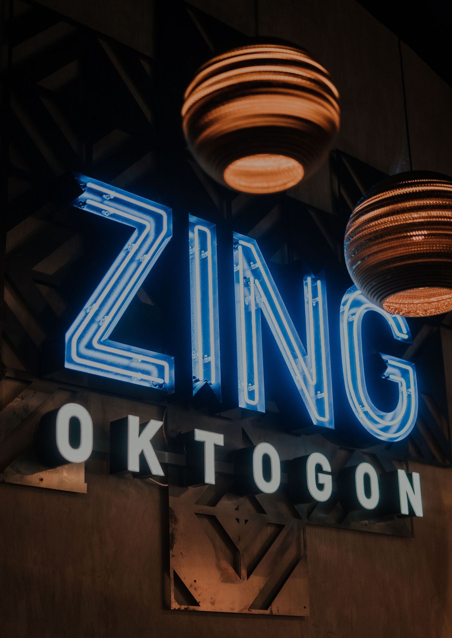 Blue neon sign reading 'ZING OKTOGON' illuminated at night with decorative hanging spherical lamps above.