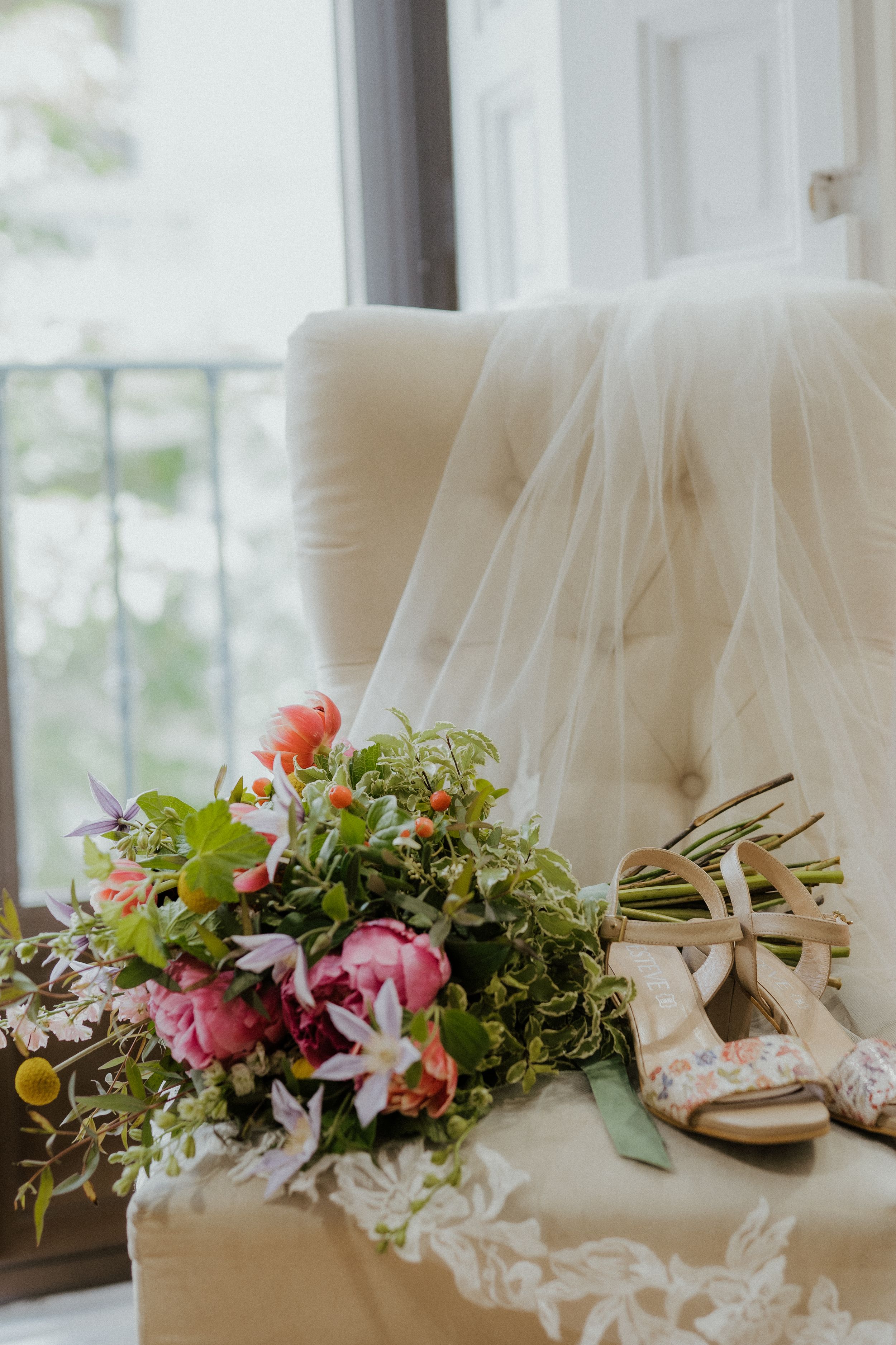 Wedding accessories with bridal veil, floral bouquet and sandals arranged on cream tufted chair by window.