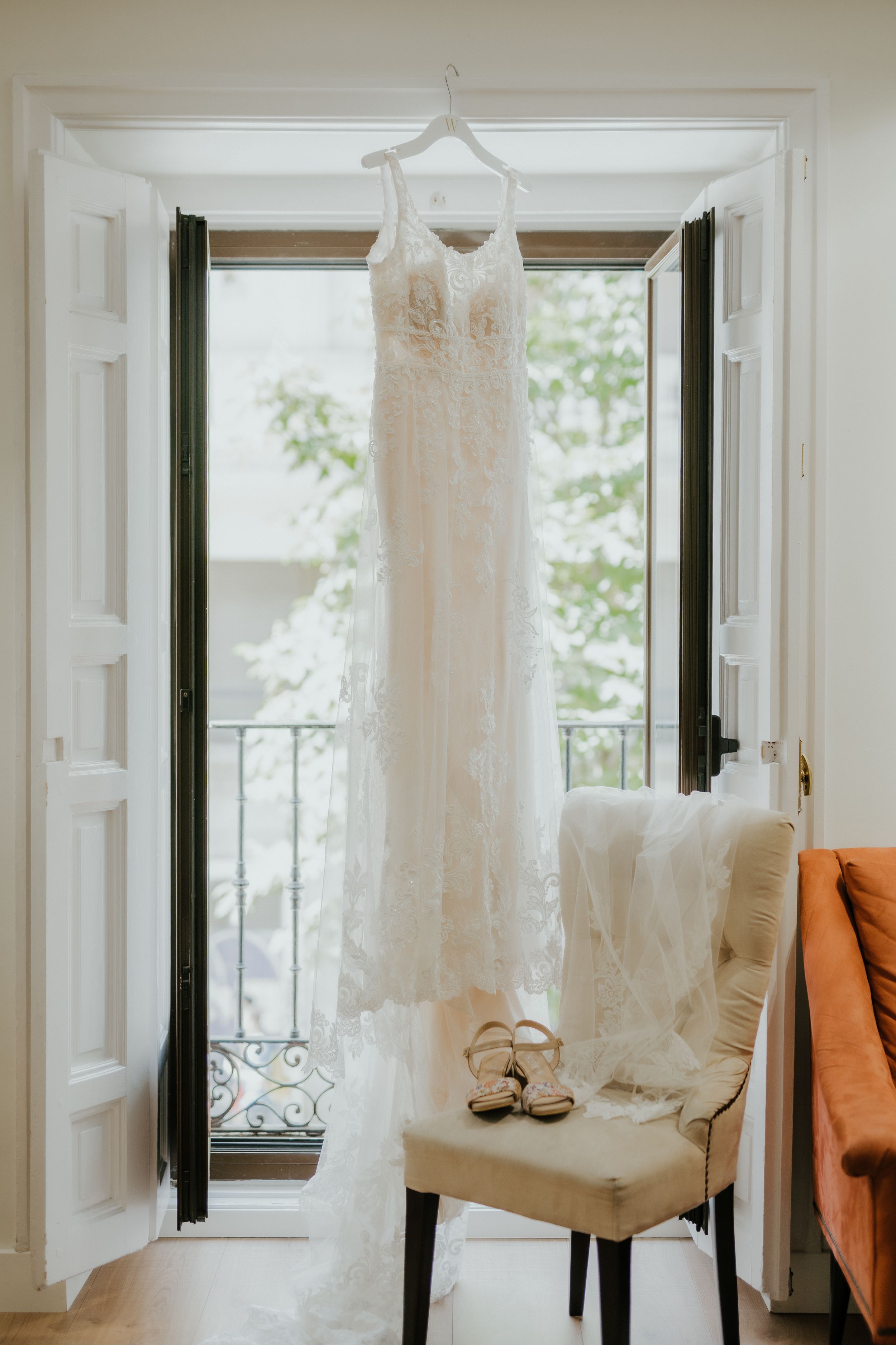 An elegant white lace wedding dress hangs in a doorway by an open window, with bridal shoes placed on a cream chair below.