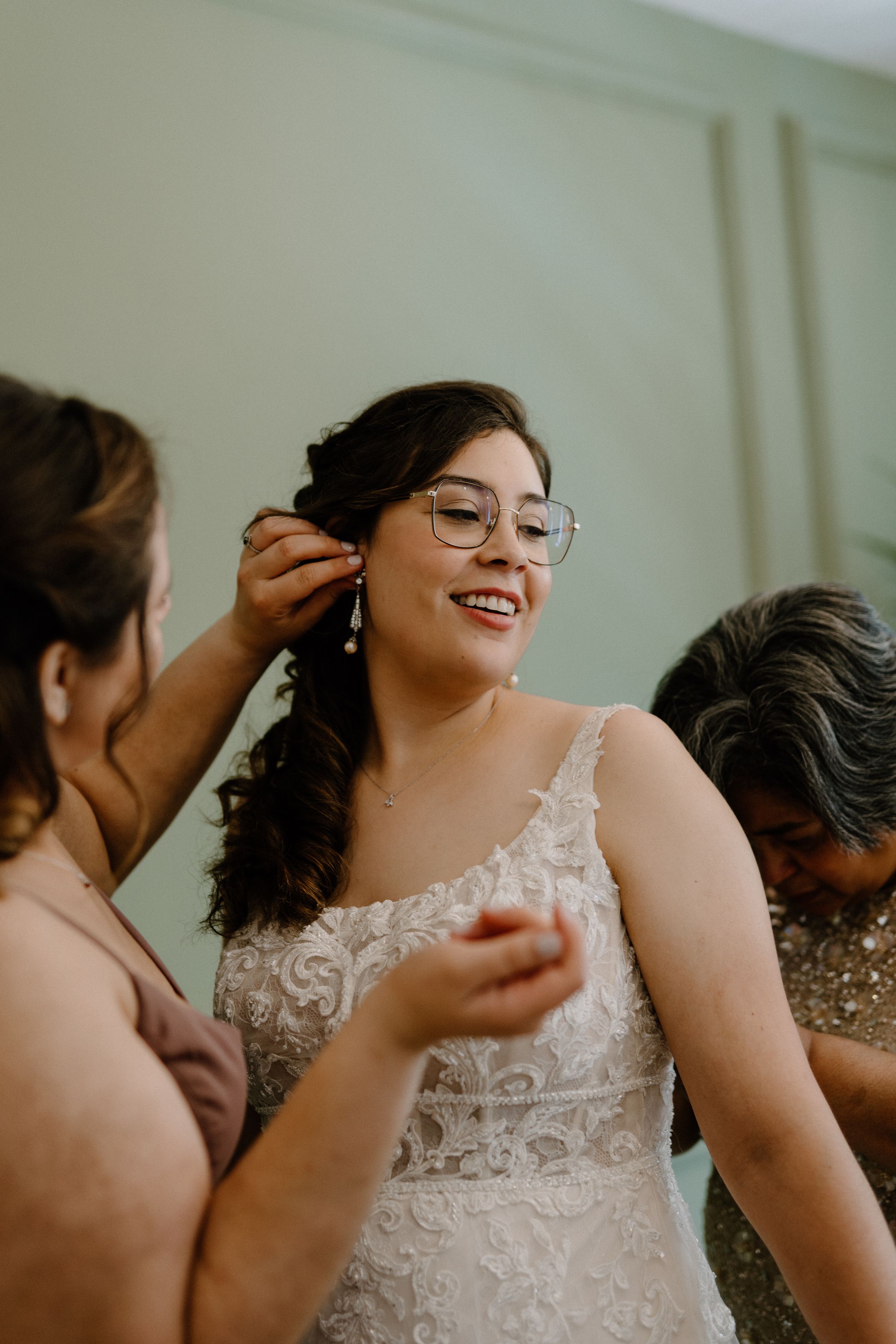 Someone wearing a white lace wedding dress smiles while having their hair styled on their wedding day.