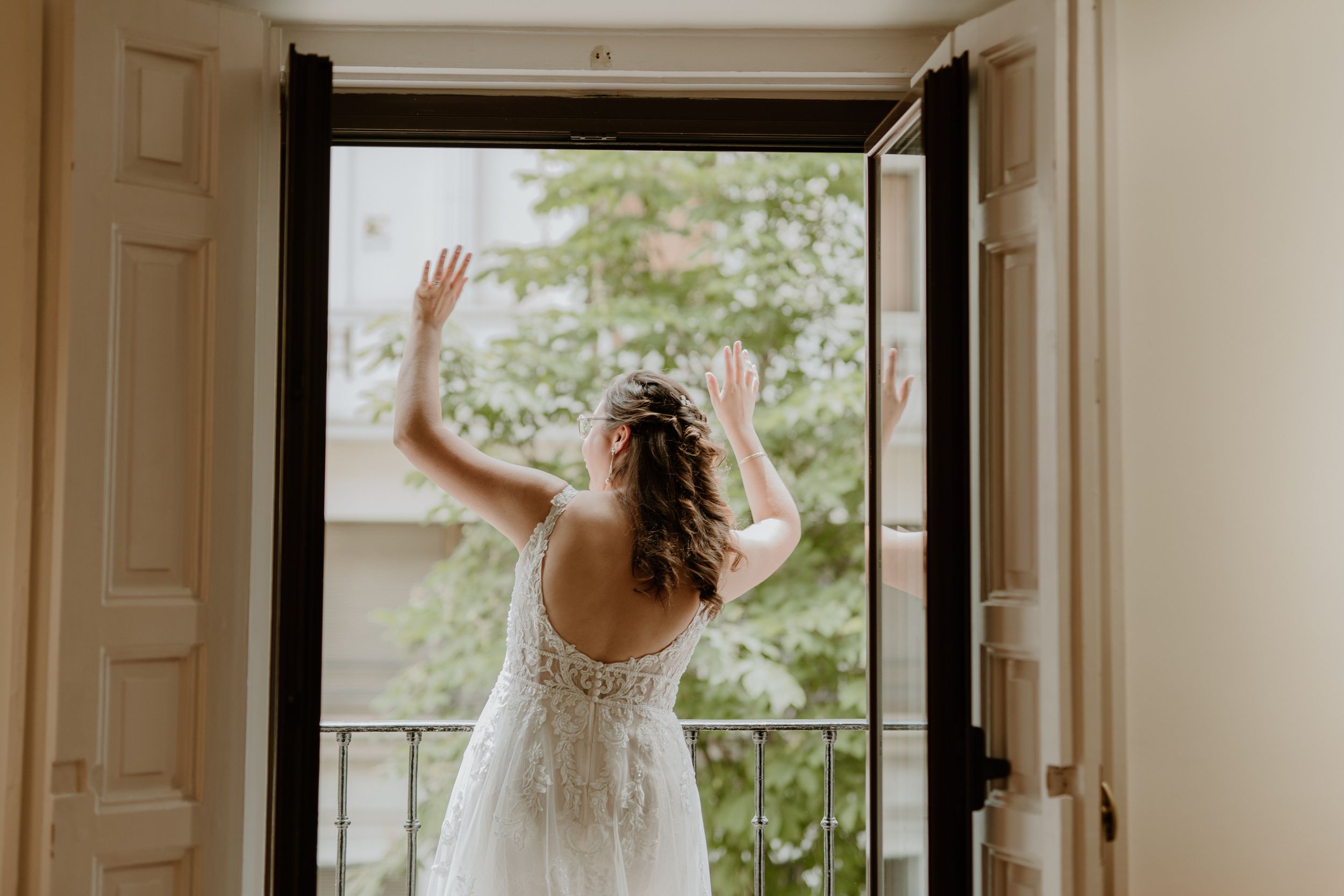 A bride in a delicate lace gown stands at a balcony doorway with her back turned, raising her hand in greeting.