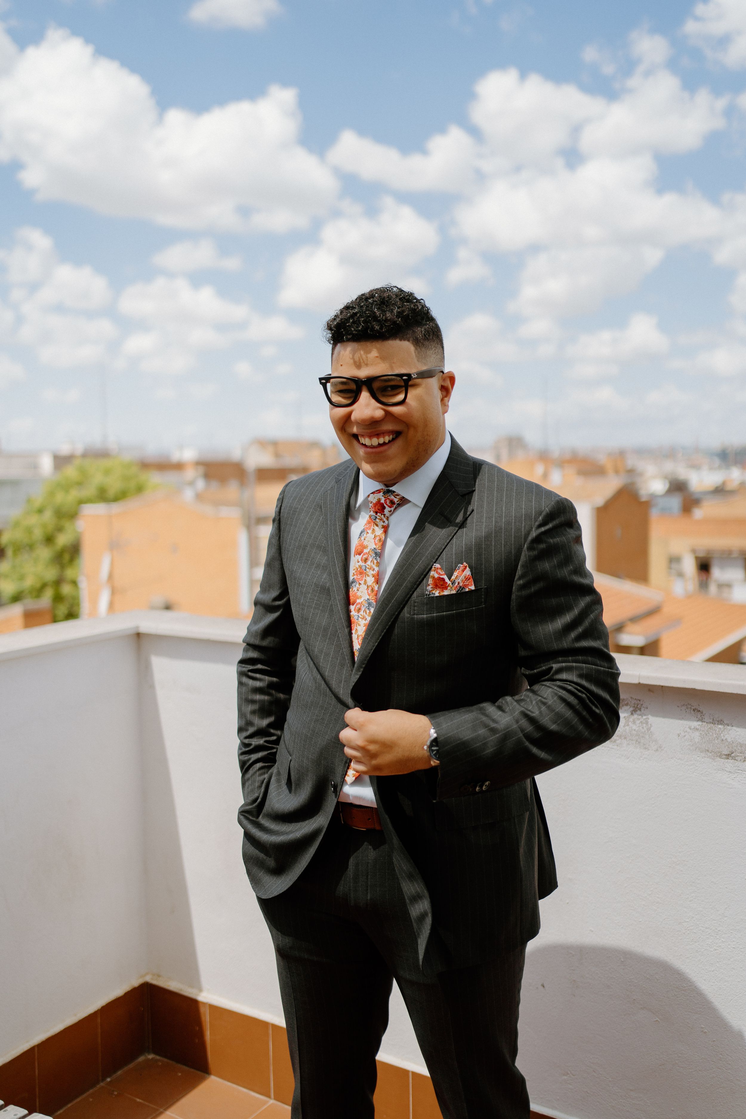 A person in a dark business suit with sunglasses and colorful tie poses on a rooftop terrace with orange city buildings behind.