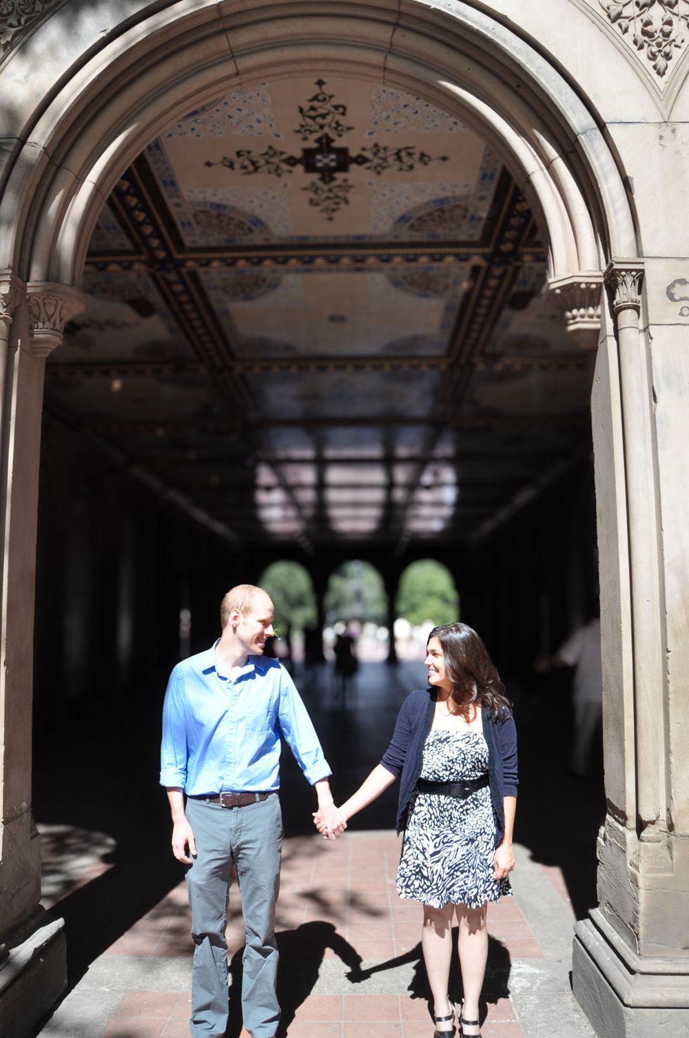 Engaged couple standing in golden sunlight with the shadowed Bethesda Terrace arches behind them in Central Park, NYC.