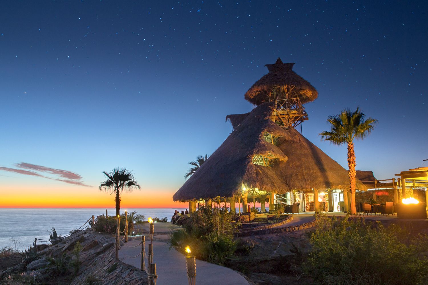 Thatched-roof resort restaurant overlooking ocean at sunset with palm trees and glowing lights. El Mirador by Guaycura.