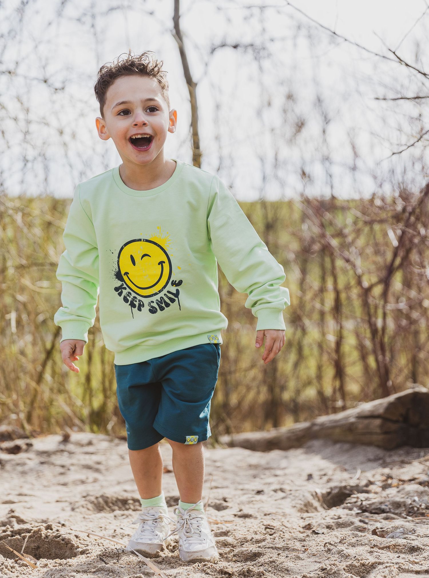 Child wearing yellow smiley face sweatshirt jumping joyfully on sandy ground.