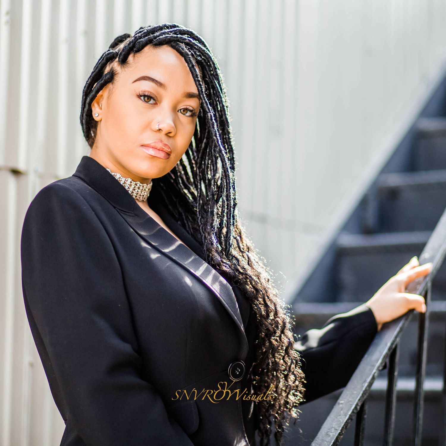 Professional portrait with elegant long braids and black blazer posing near stair railing with natural lighting.