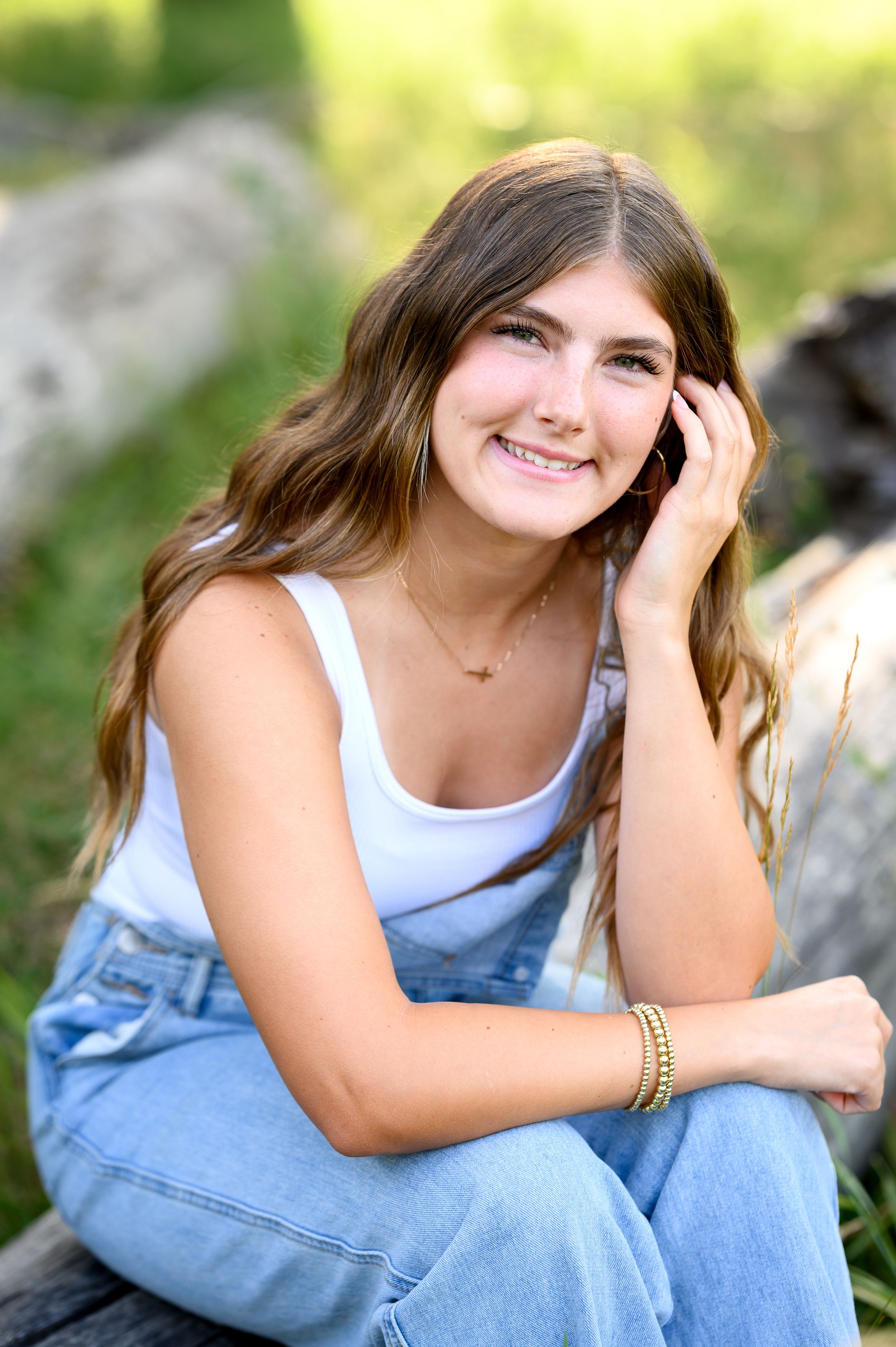 A model in overalls and white tank top poses casually against a rustic outdoor setting with natural bokeh background. Wallowa Lake, Joseph,