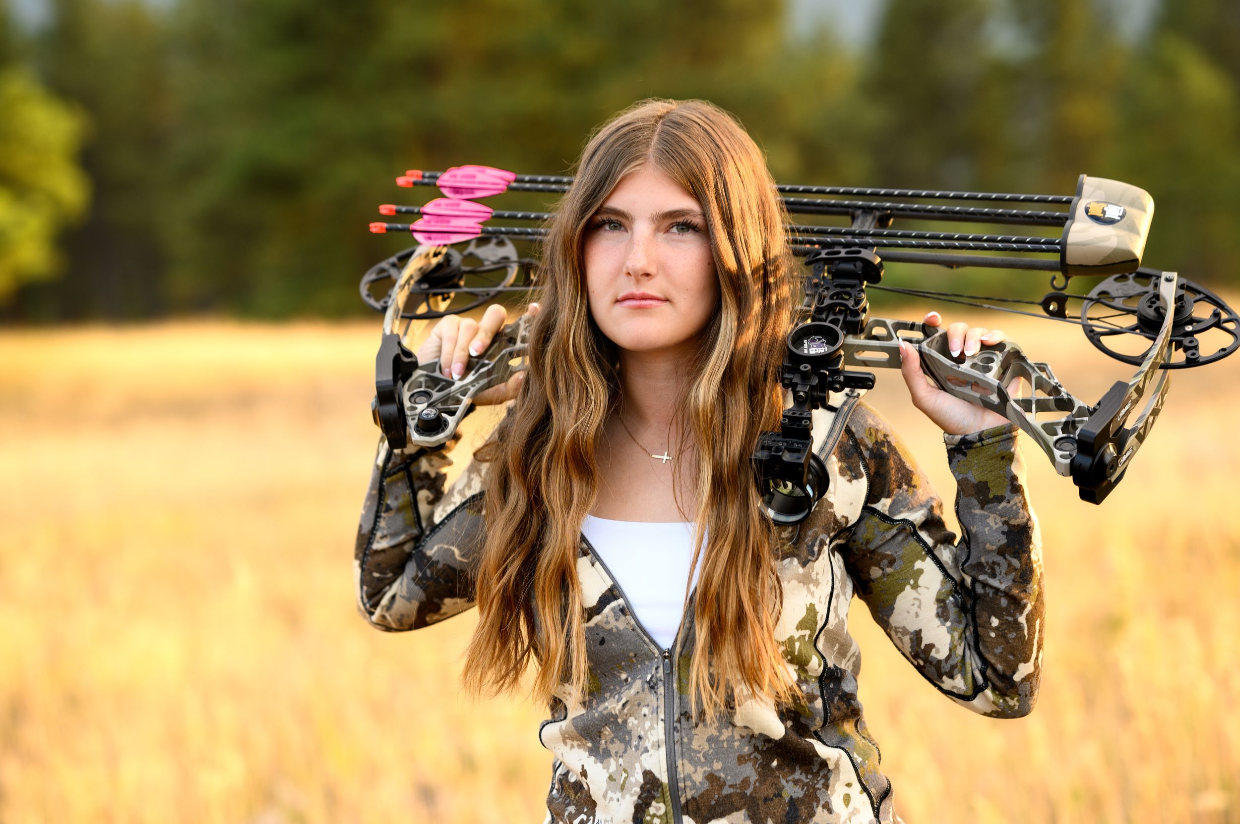 Archer holding two camouflage compound bows in golden wheat field.