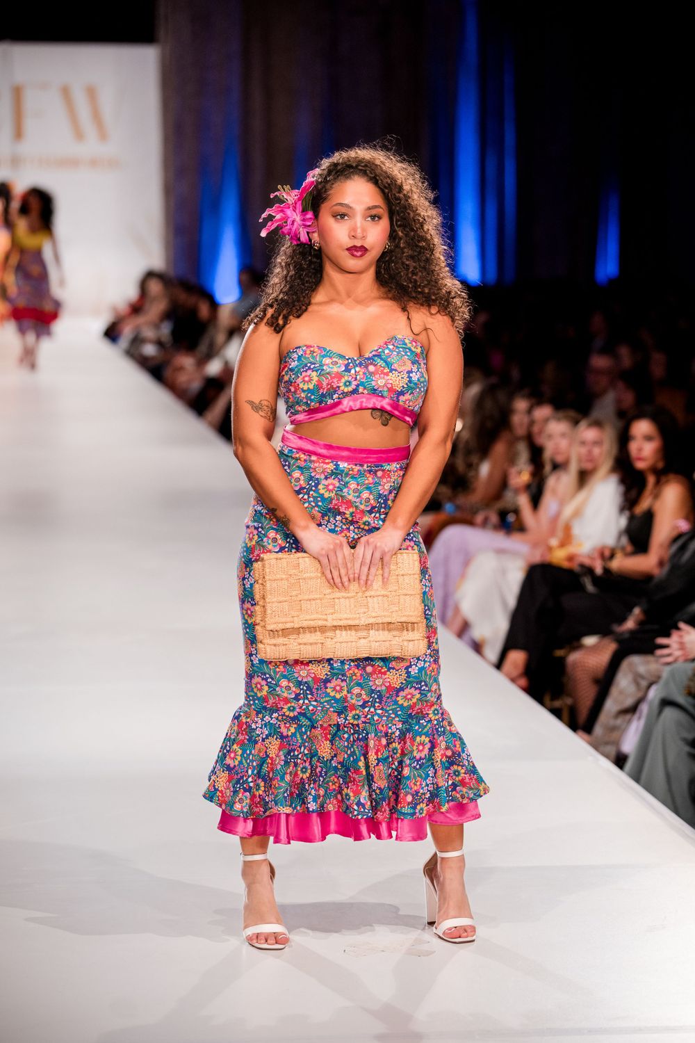 Floral print two-piece outfit displayed on fashion runway with audience in background.