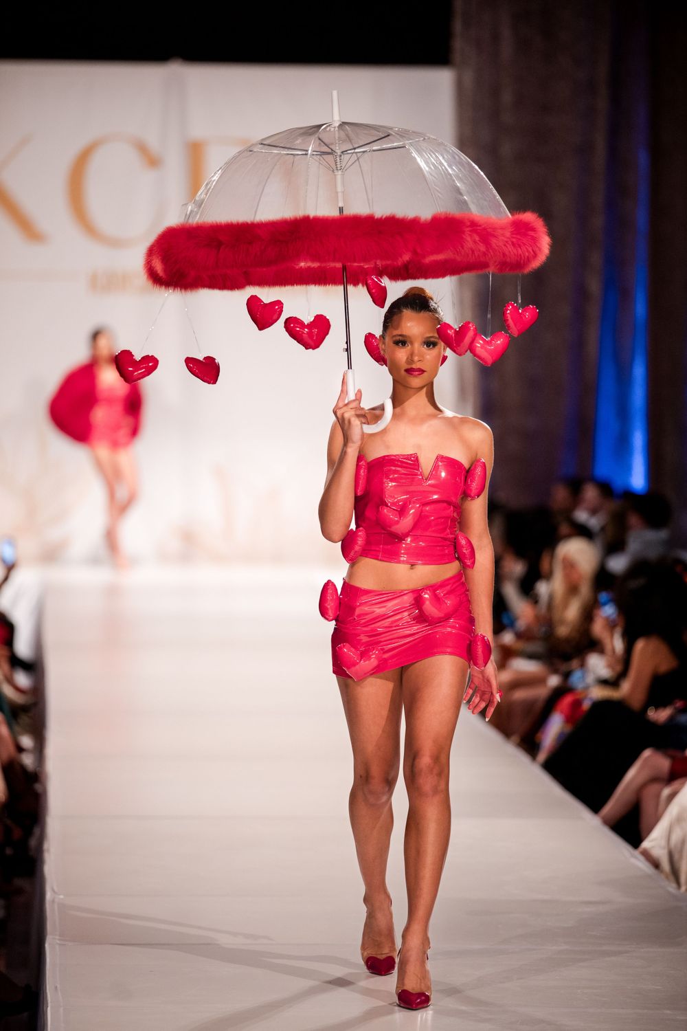Fashion model walks runway in vibrant red ensemble with heart-adorned umbrella at fashion show.