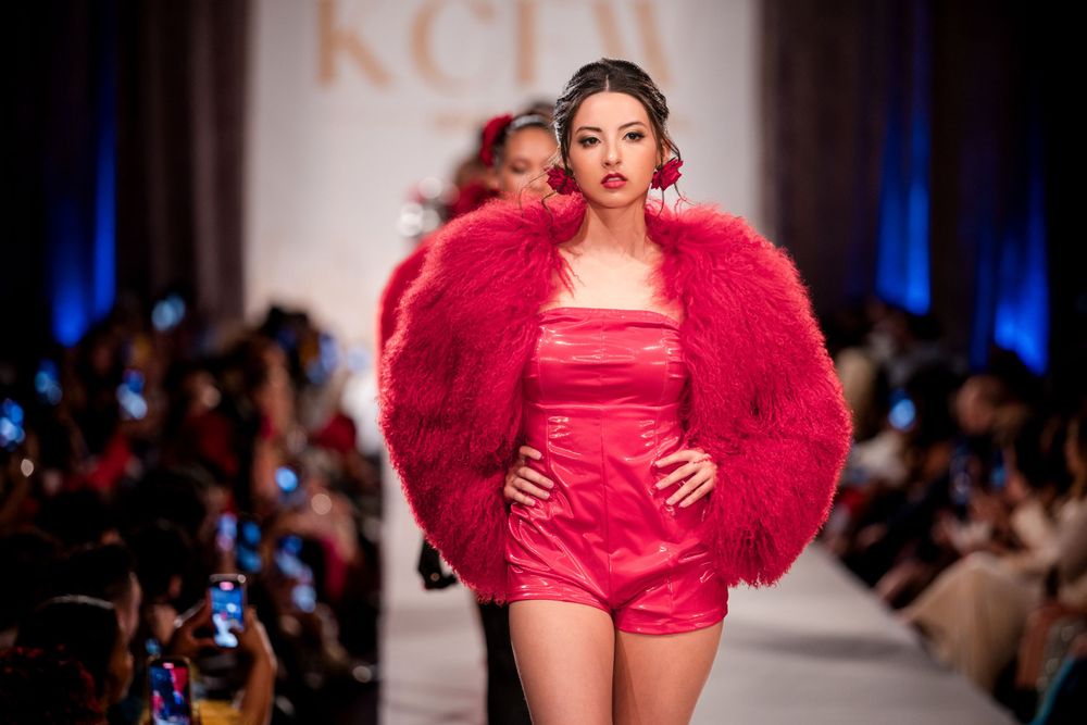 Model showcases bright red couture dress with dramatic ruffles on illuminated catwalk.