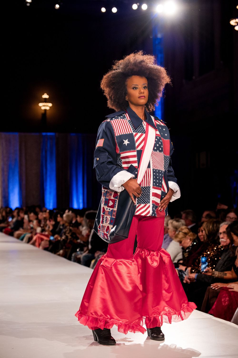 Runway model showcases patriotic American flag jacket with red ruffled skirt on fashion catwalk.