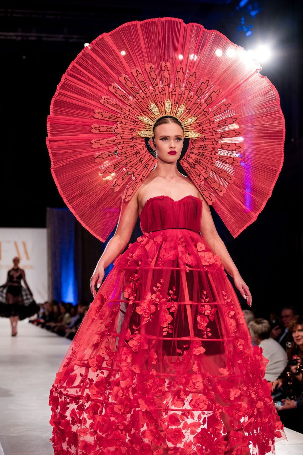 Dramatic red fan-shaped couture gown with elaborate details displayed on illuminated runway.