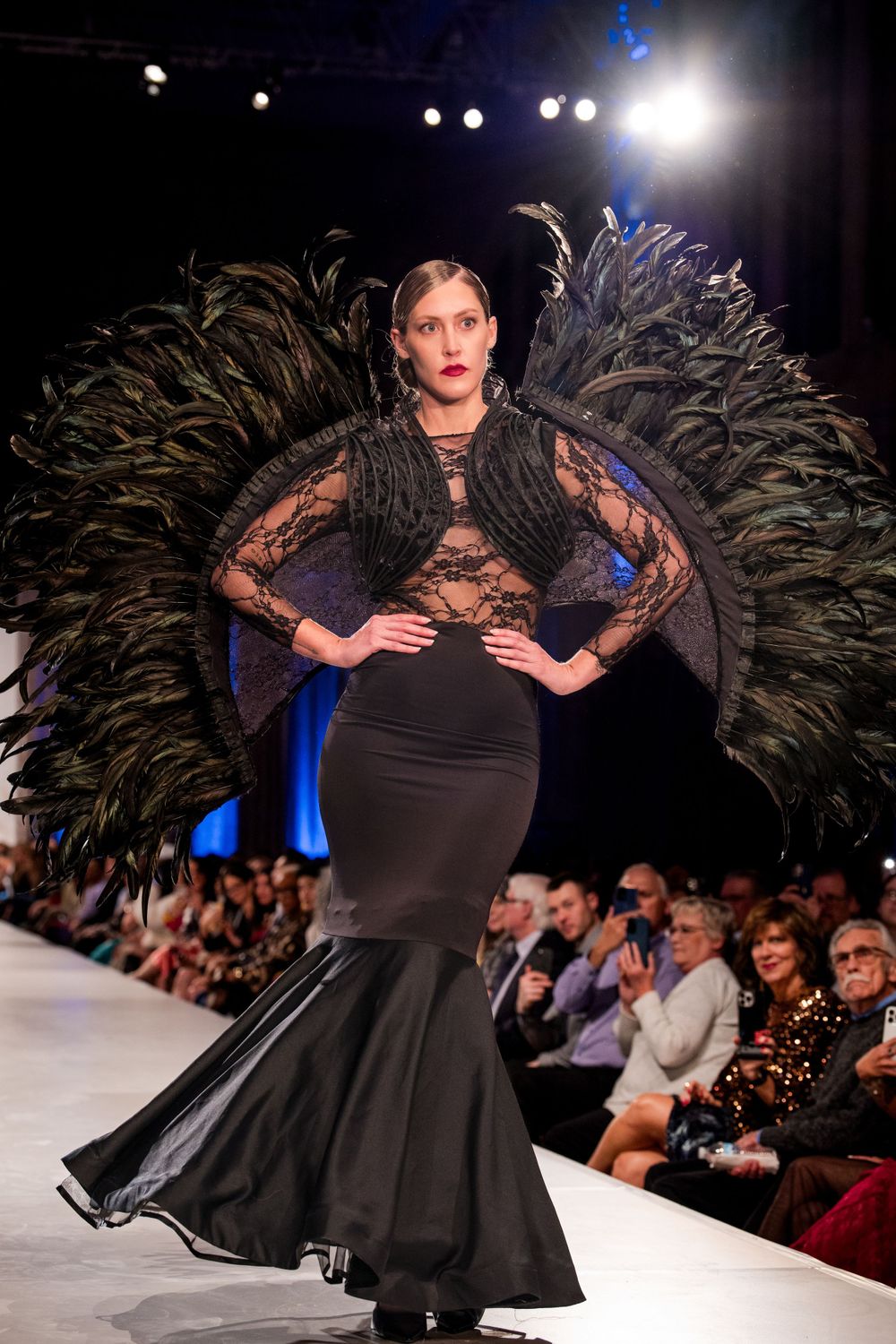 Model presents elegant black dress with dramatic feather details on fashion runway.