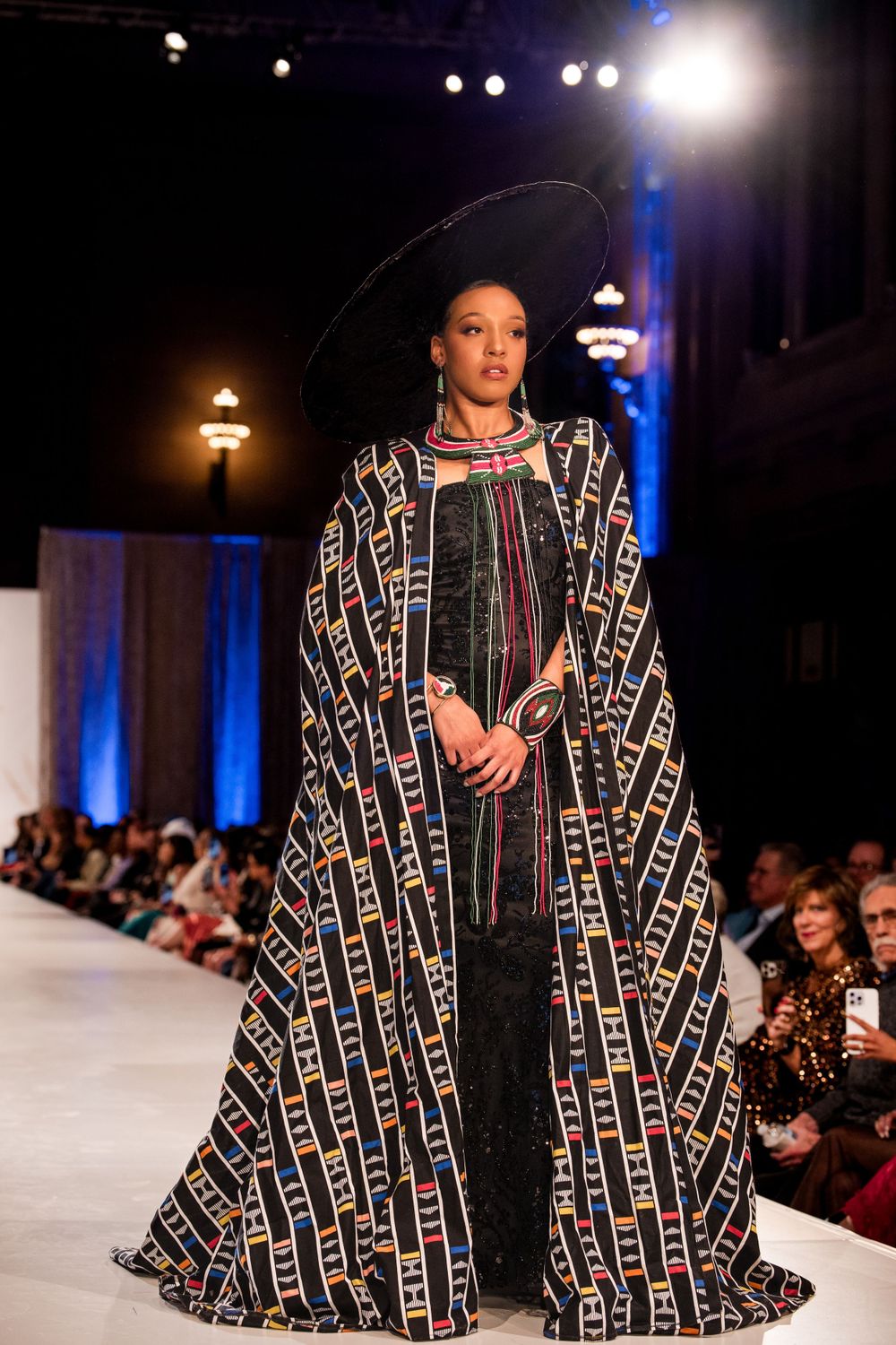Geometric patterned cape coat in black and gold shown on illuminated fashion runway.
