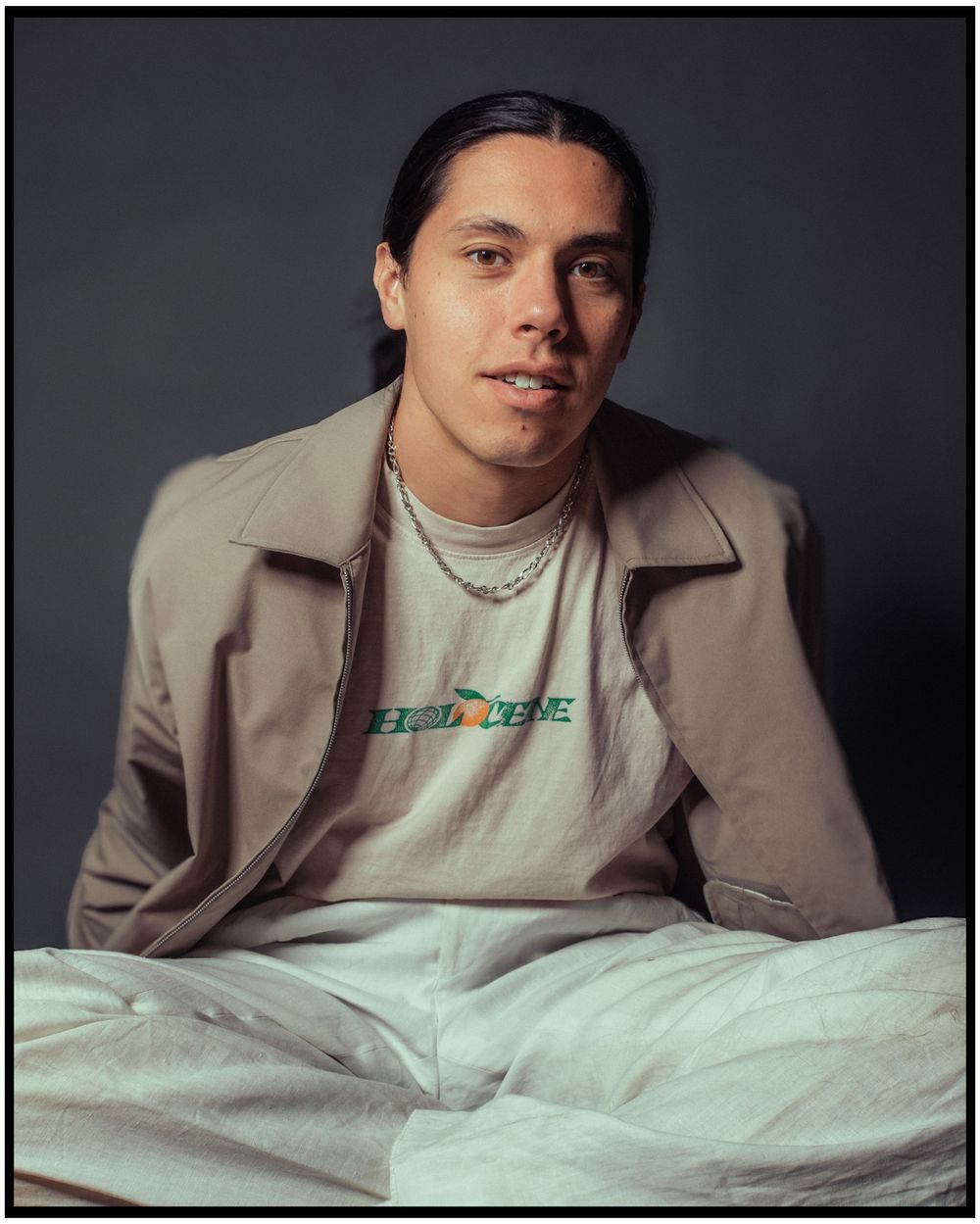 Professional studio portrait of person wearing a beige jacket and graphic t-shirt against dark background.