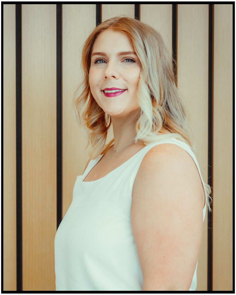 Professional portrait photograph of someone with wavy blonde hair wearing a white sleeveless top against a striped background.