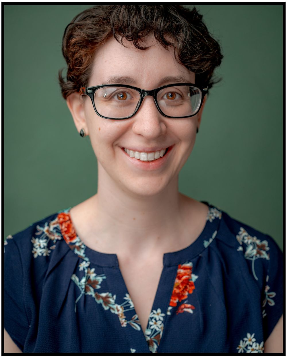 Professional headshot of someone wearing glasses and a navy floral print top against a green background.