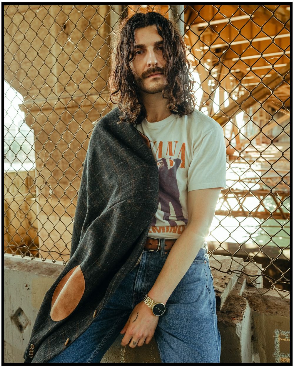 Person in a vintage t-shirt and jeans posing against a chain-link fence with long wavy hair.