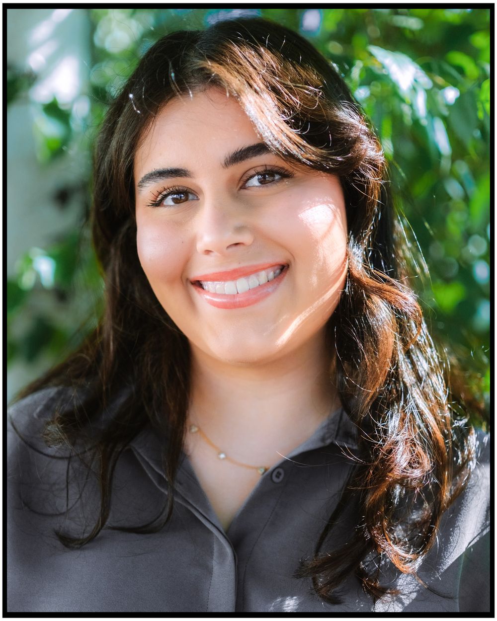 Portrait of someone wearing a dark shirt smiling outdoors with sunlit foliage in the background.