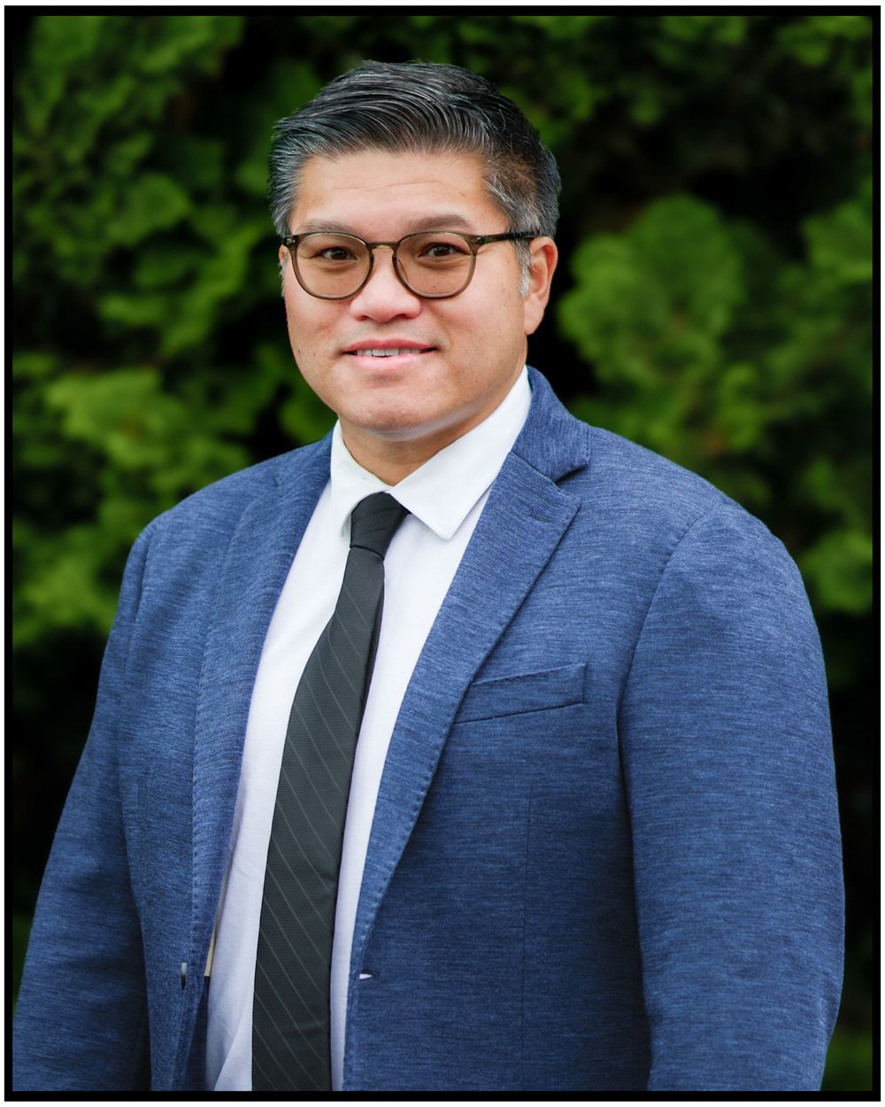 Professional headshot portrait of someone in a navy suit and glasses against a green outdoor background.