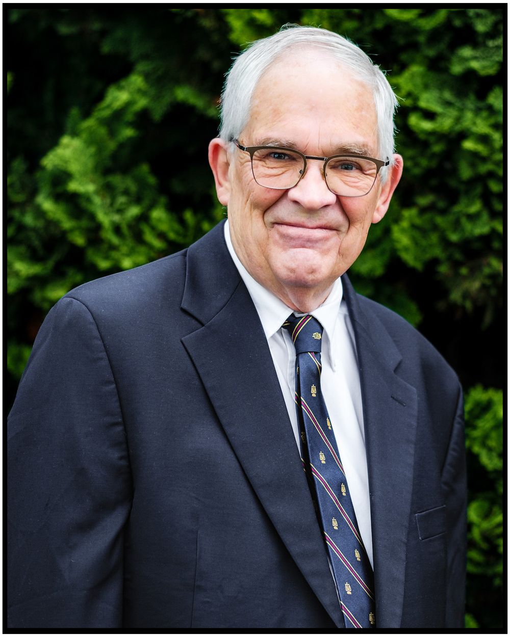 Professional portrait of someone in dark suit and tie standing in front of green foliage.