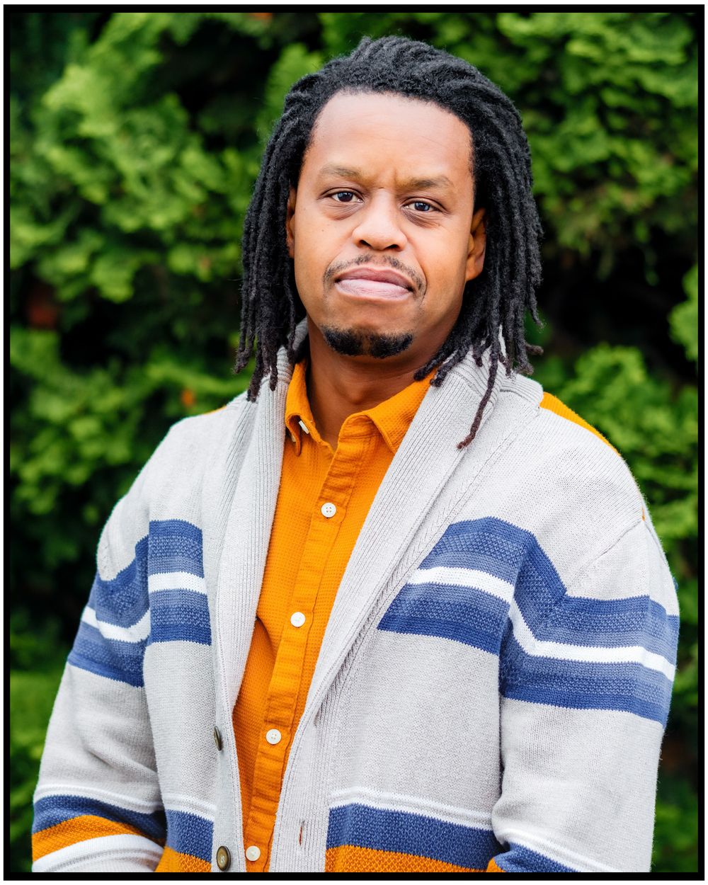 Portrait of a person with locs wearing an orange button-down shirt and gray cardigan with blue stripes against green foliage.