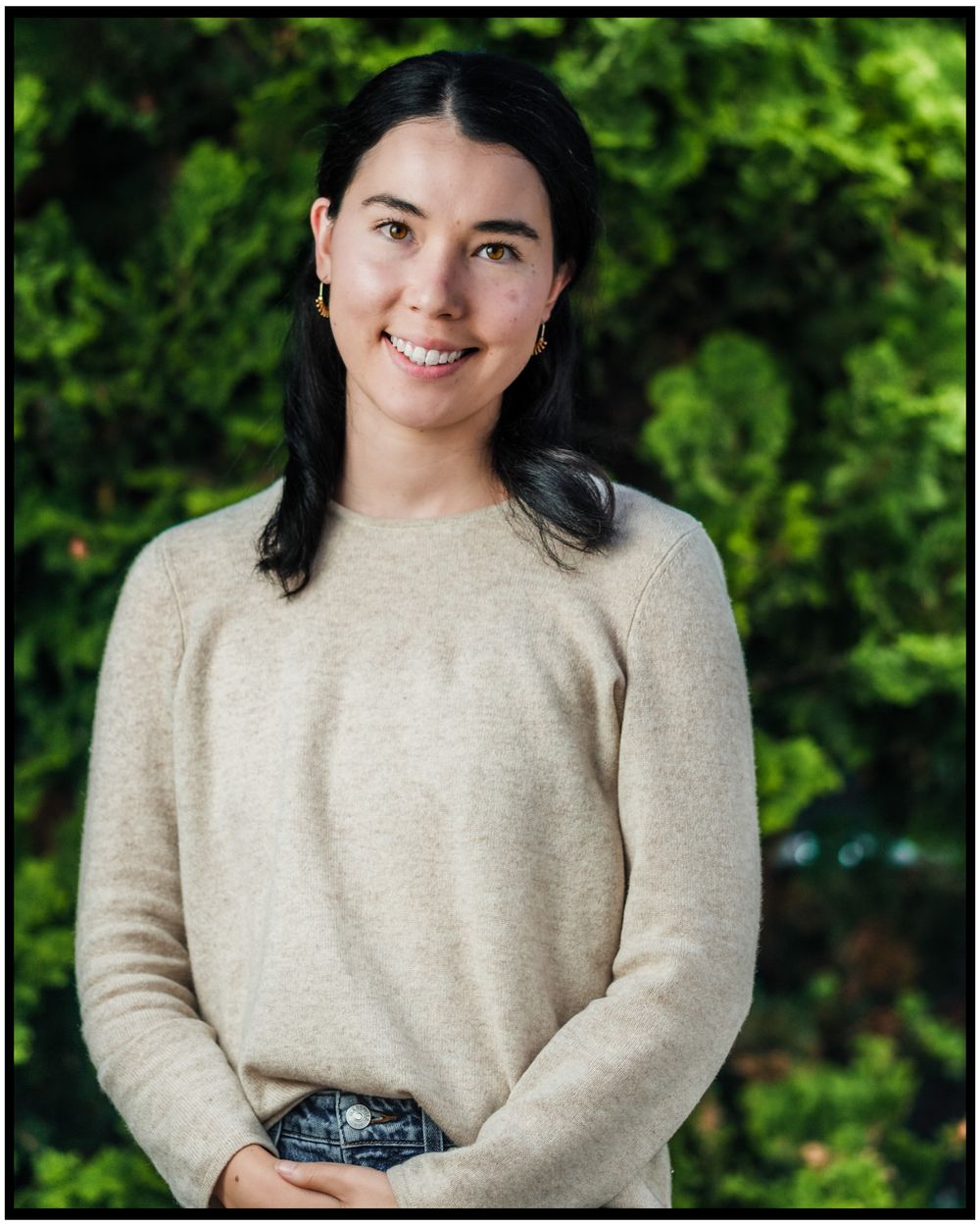 Outdoor portrait of person with dark hair wearing cream sweater against lush green background.