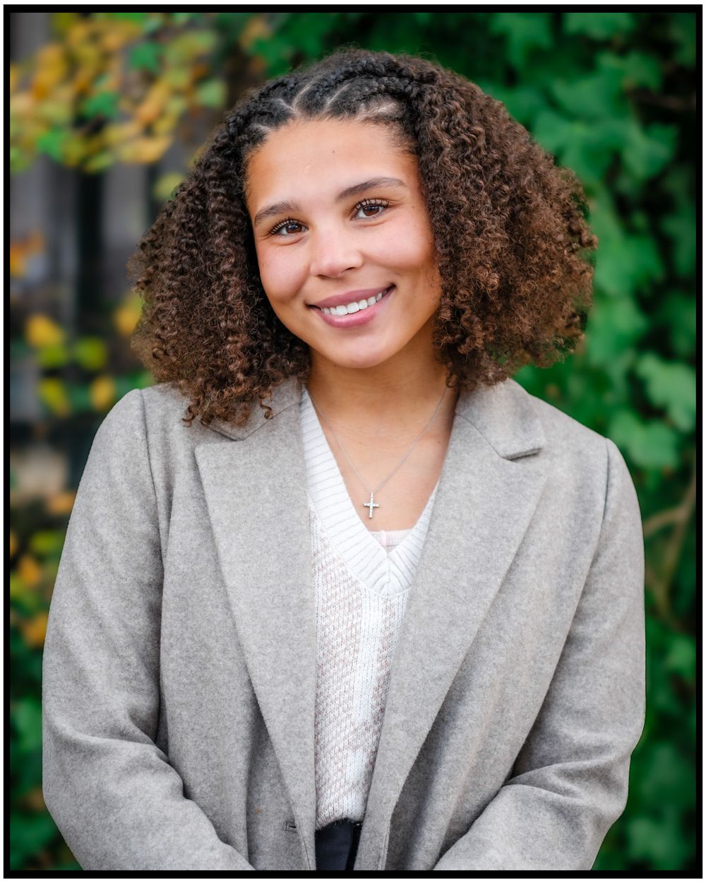 Professional headshot of a person with curly hair wearing a gray blazer and white sweater against a blurred green background.