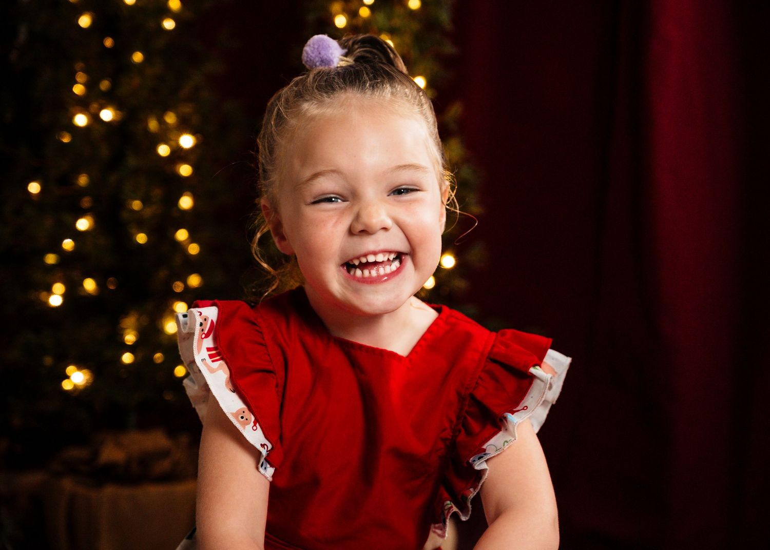 Happy child in red holiday dress laughing with Christmas lights in background.