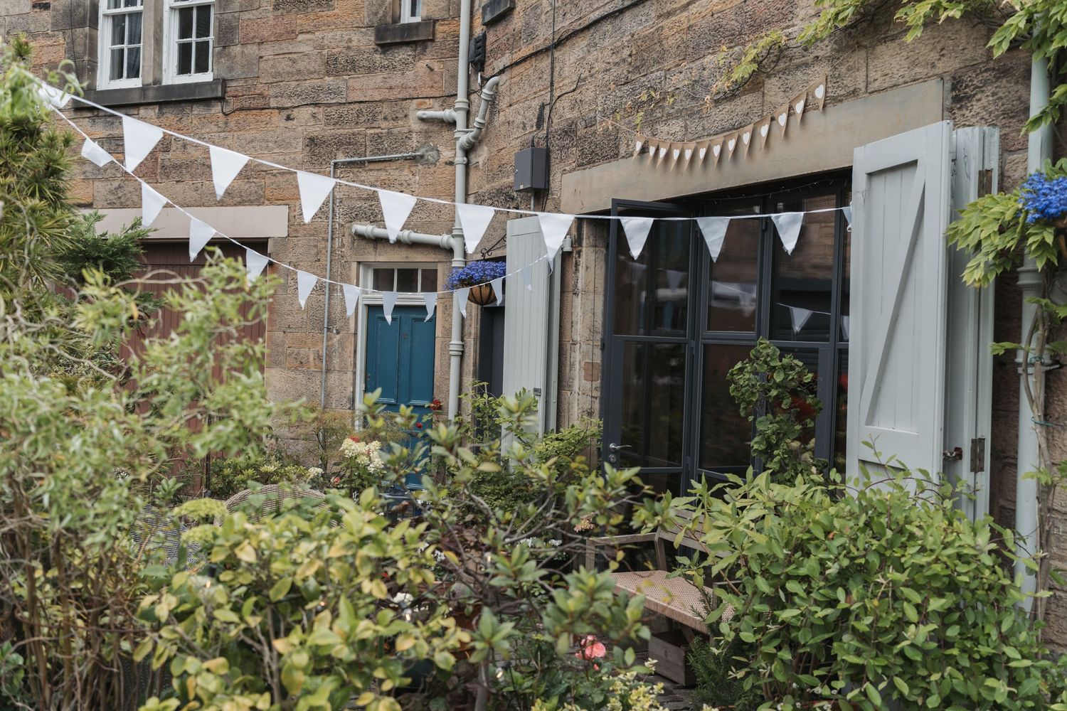 White bunting decorates the exterior of a charming stone building with blue door and garden shrubs in front.