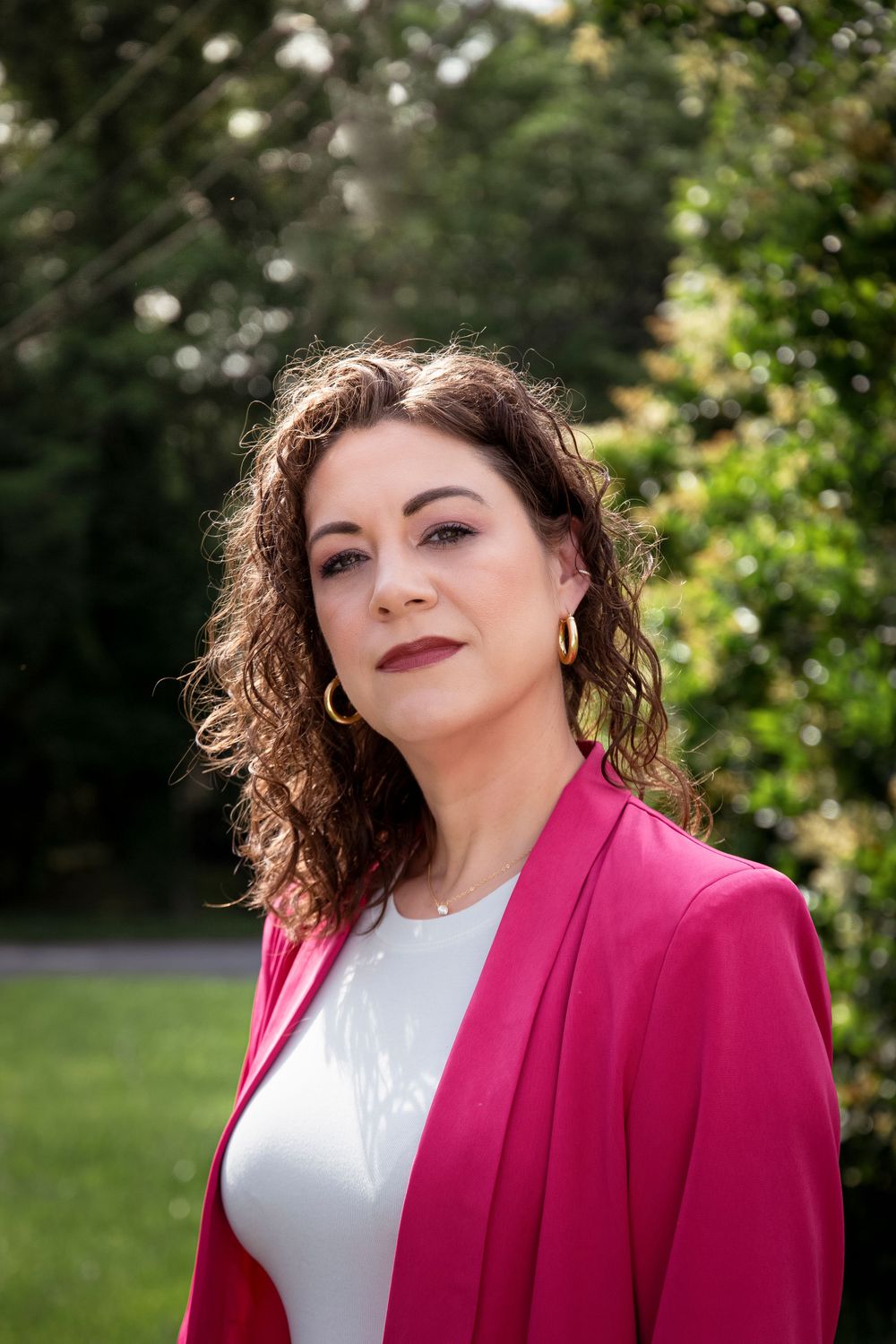 Professional portrait of figure in vibrant pink blazer and white top against outdoor garden background.