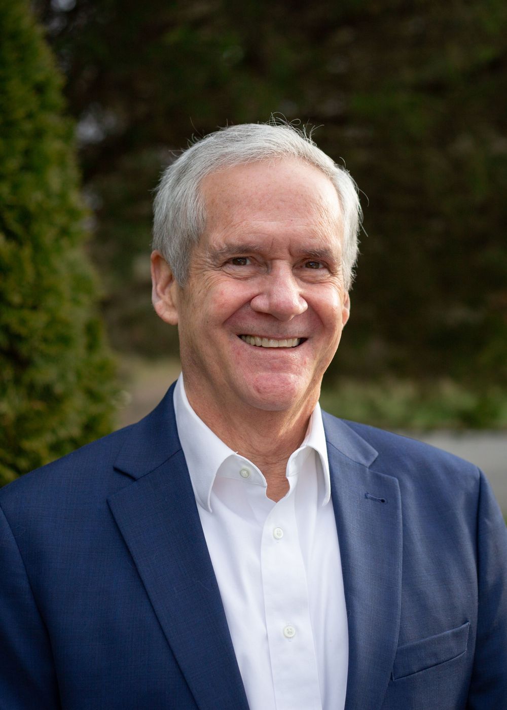 Professional headshot of senior man in navy blazer smiling outdoors.